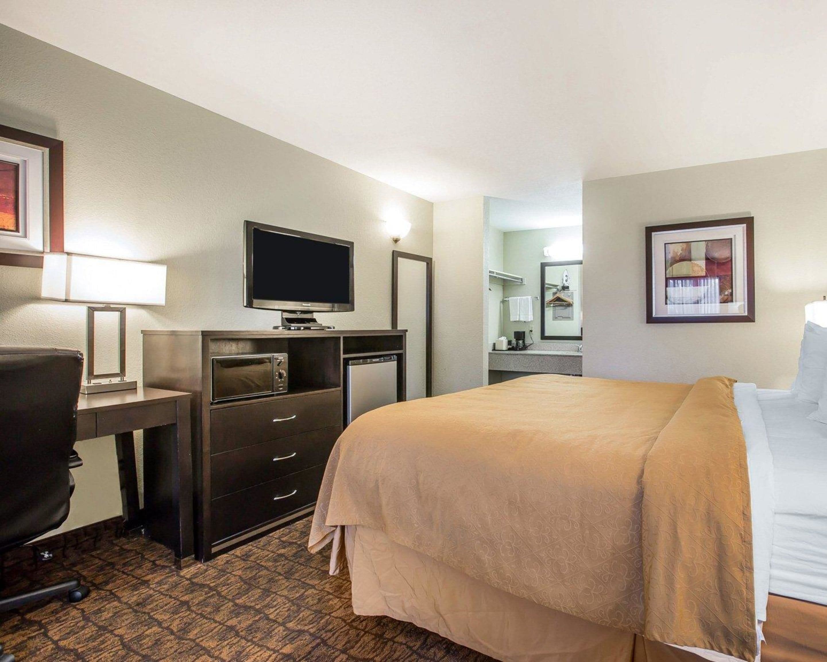 Habitación de hotel con cama, escritorio con televisor, silla, cómoda y baño visible a través de una puerta abierta.