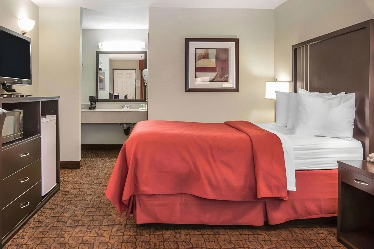 A hotel room with a bed, a desk, a television, a mirror, and a bathroom visible in the background.