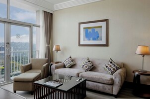 Habitación de hotel moderna con sofá, sillón y mesa de centro, un ventanal con vistas a la ciudad y un cuadro abstracto enmarcado en la pared.
