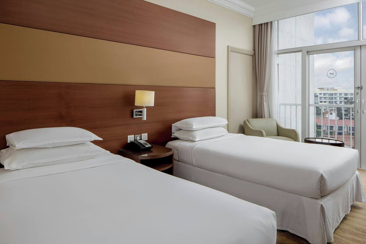 A hotel room with two beds, a nightstand with a lamp, a chair, and a view of buildings through a large window.