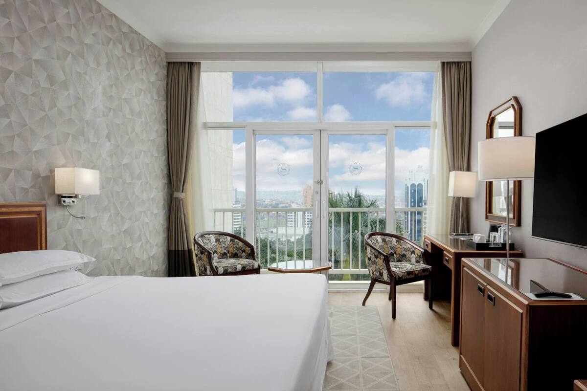 A hotel room with a bed, two chairs, a desk, a TV, and a balcony with a city view.