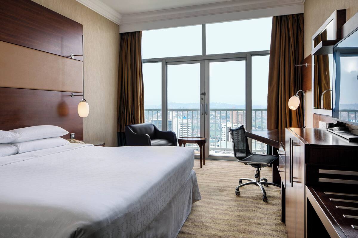 A hotel room with a large bed, a desk, a chair, and a TV. The room has a large window with a view of the city.