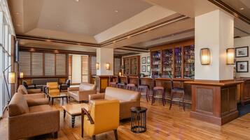 A spacious lobby with leather sofas, wooden flooring, a bar area with stools, and wall-mounted lighting.