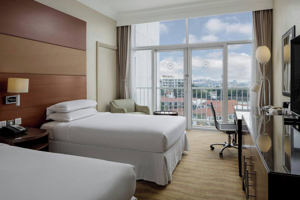 A hotel room with two beds, a desk, a chair, a TV, and a large window with a city view.