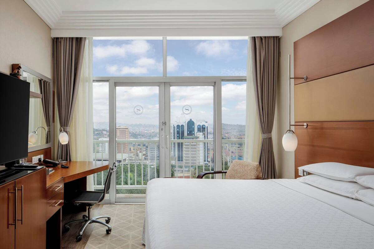 A hotel room with a large bed, a desk with a computer, a chair, and a view of the cityscape through large windows.