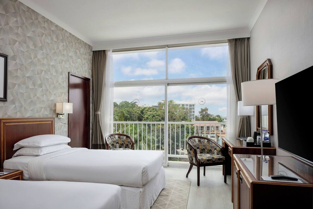 A hotel room with two beds, a desk with a TV, a chair, and a balcony with a view of buildings and trees.