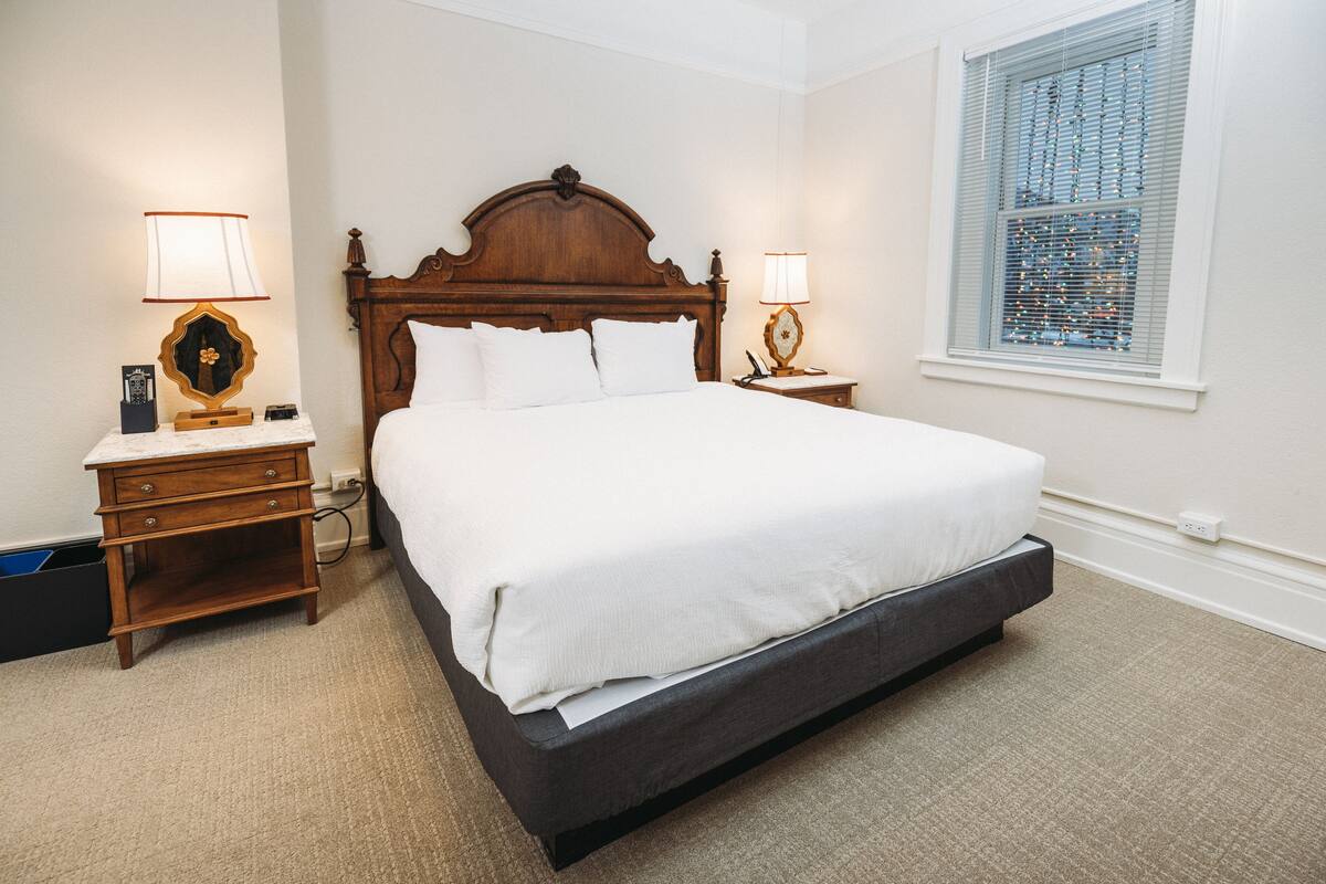 A bedroom with a large bed, wooden headboard, two bedside tables with lamps, a nightstand, and a window with blinds.