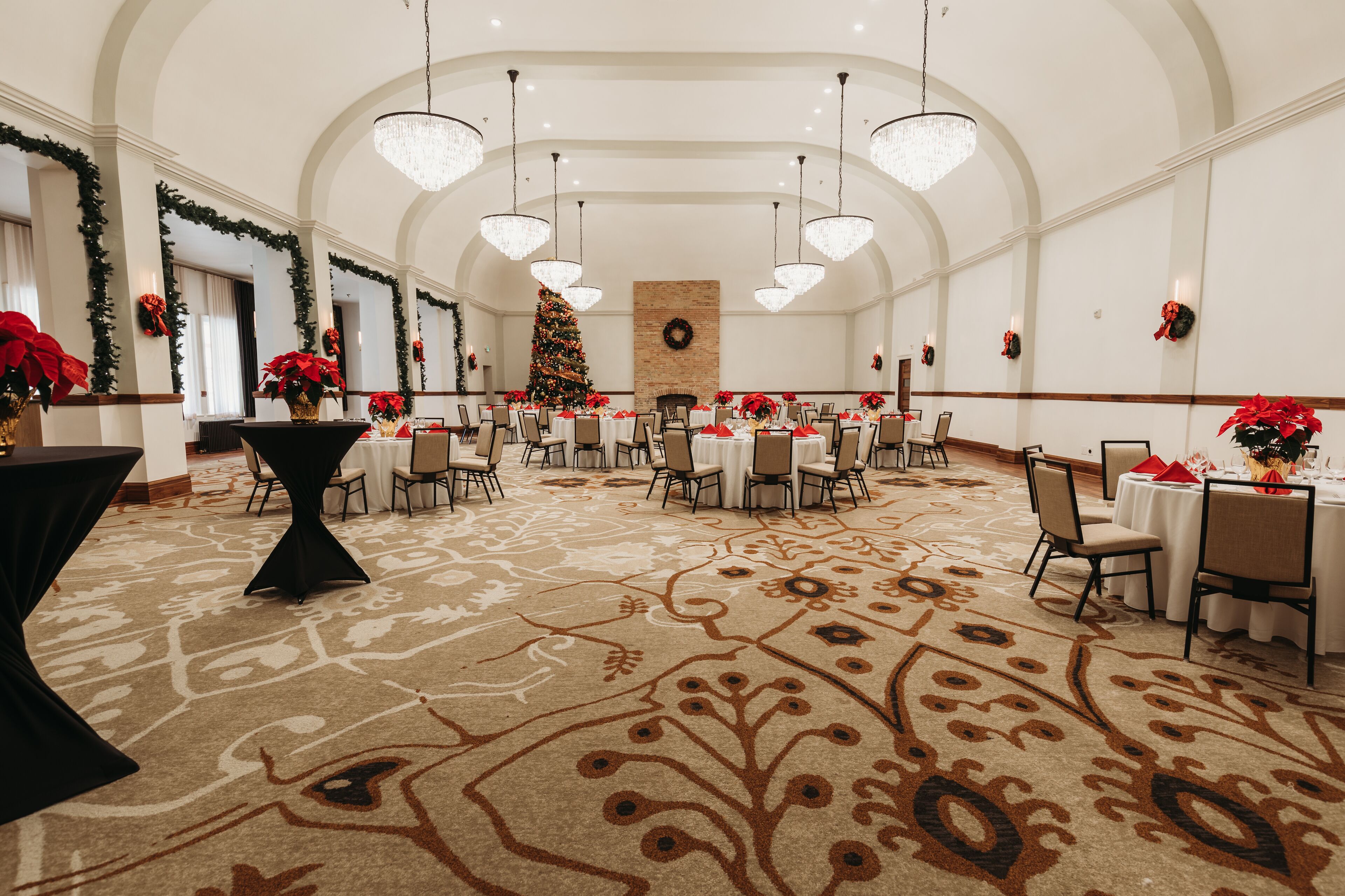 Una gran sala de banquet decorada amb elegància, amb alts sostres, lluminàries penjants i un arbre de Nadal al centre. Les taules tenen estovat blanc i elements decoratius vermells.
