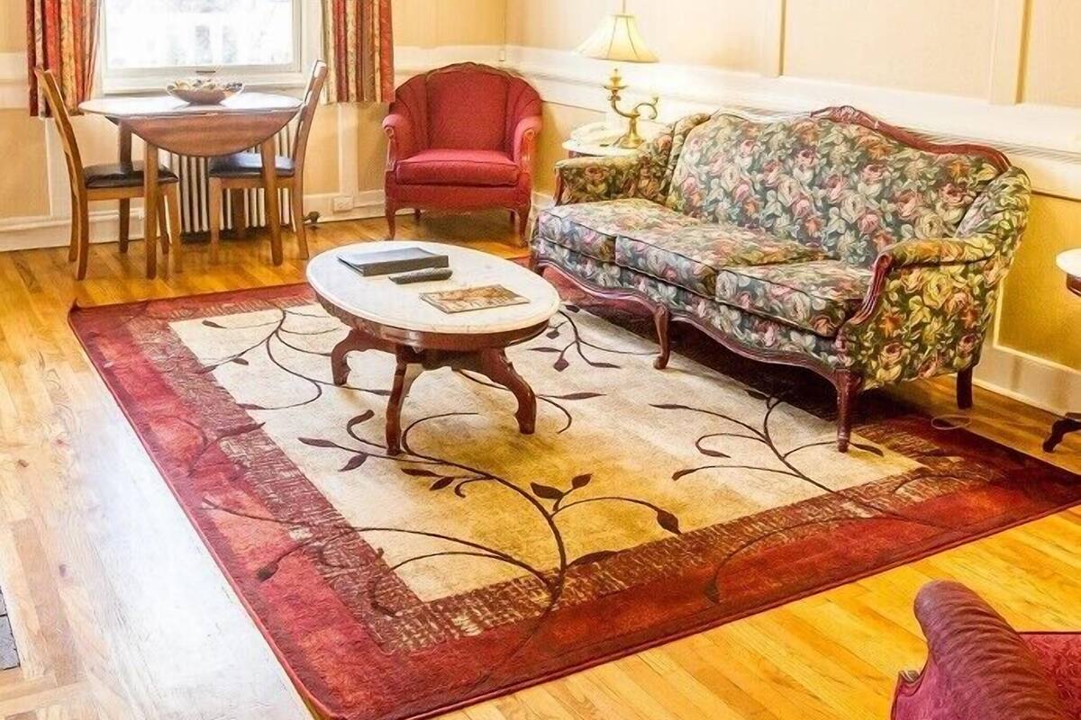 A living room with a floral sofa, a round coffee table, a red armchair, a dining table with chairs, and a window with curtains.