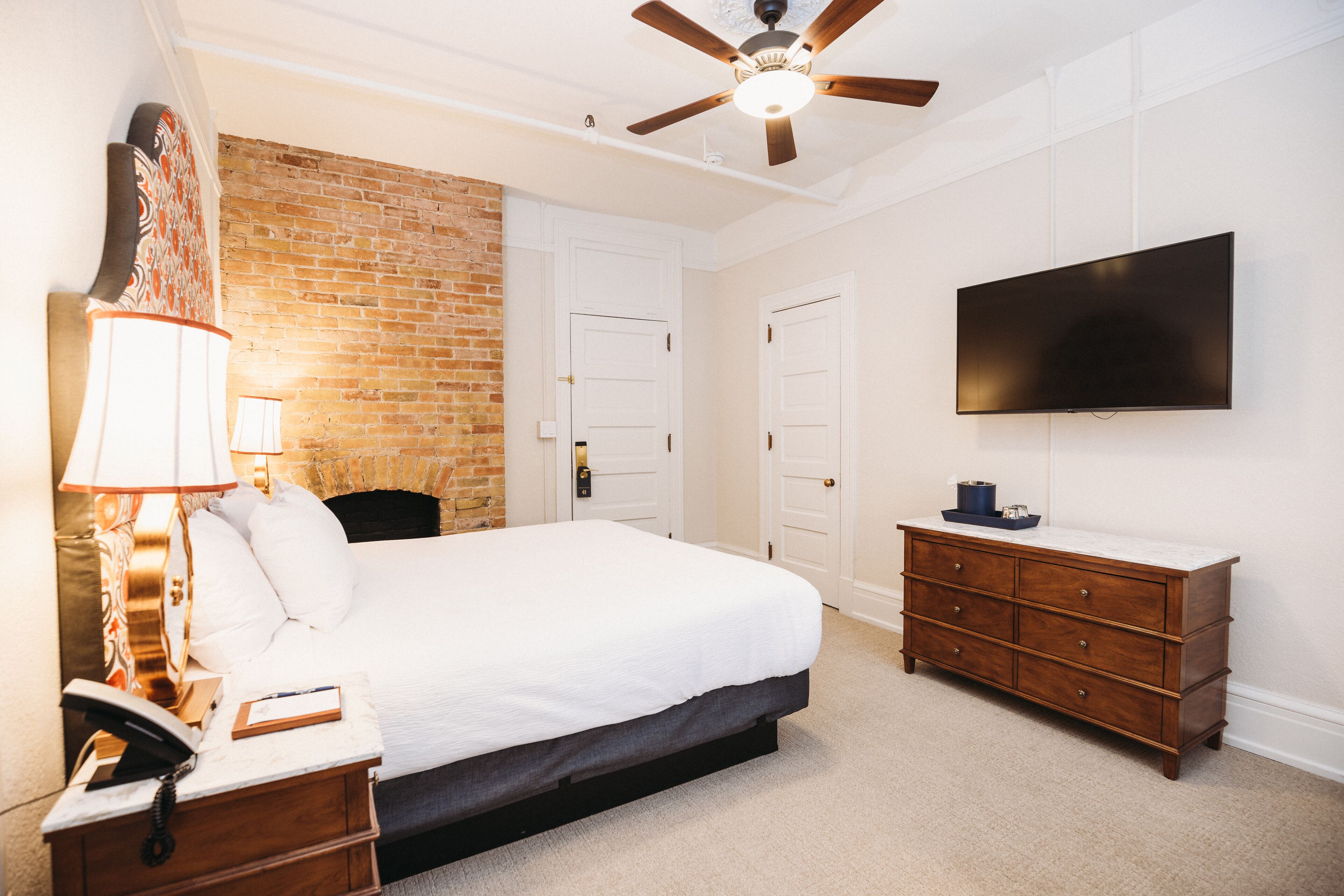 A bedroom with a brick wall, a bed, a dresser, a TV, and a ceiling fan.