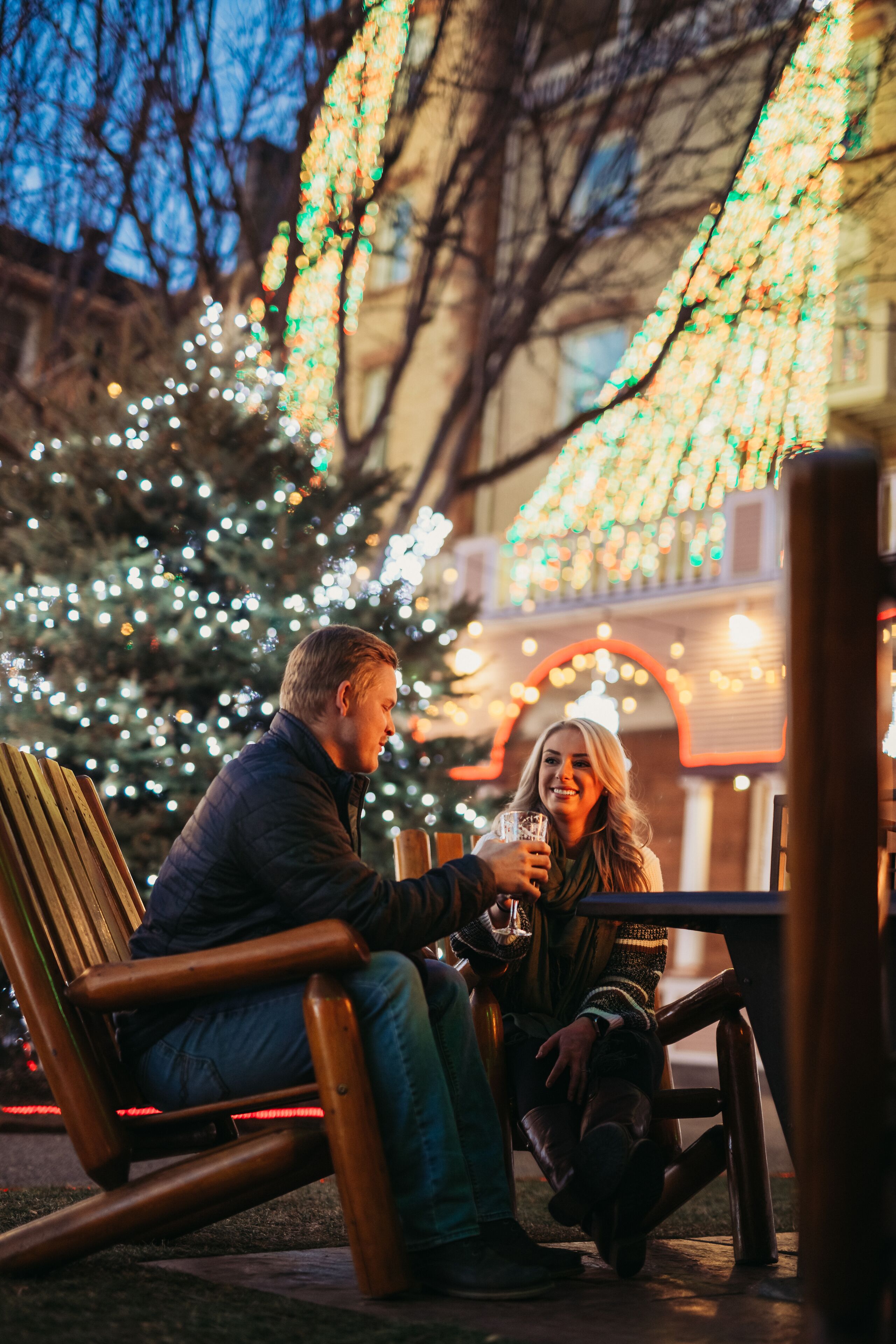 A couple seated at an outdoor table with a Christmas tree and lights in the background.