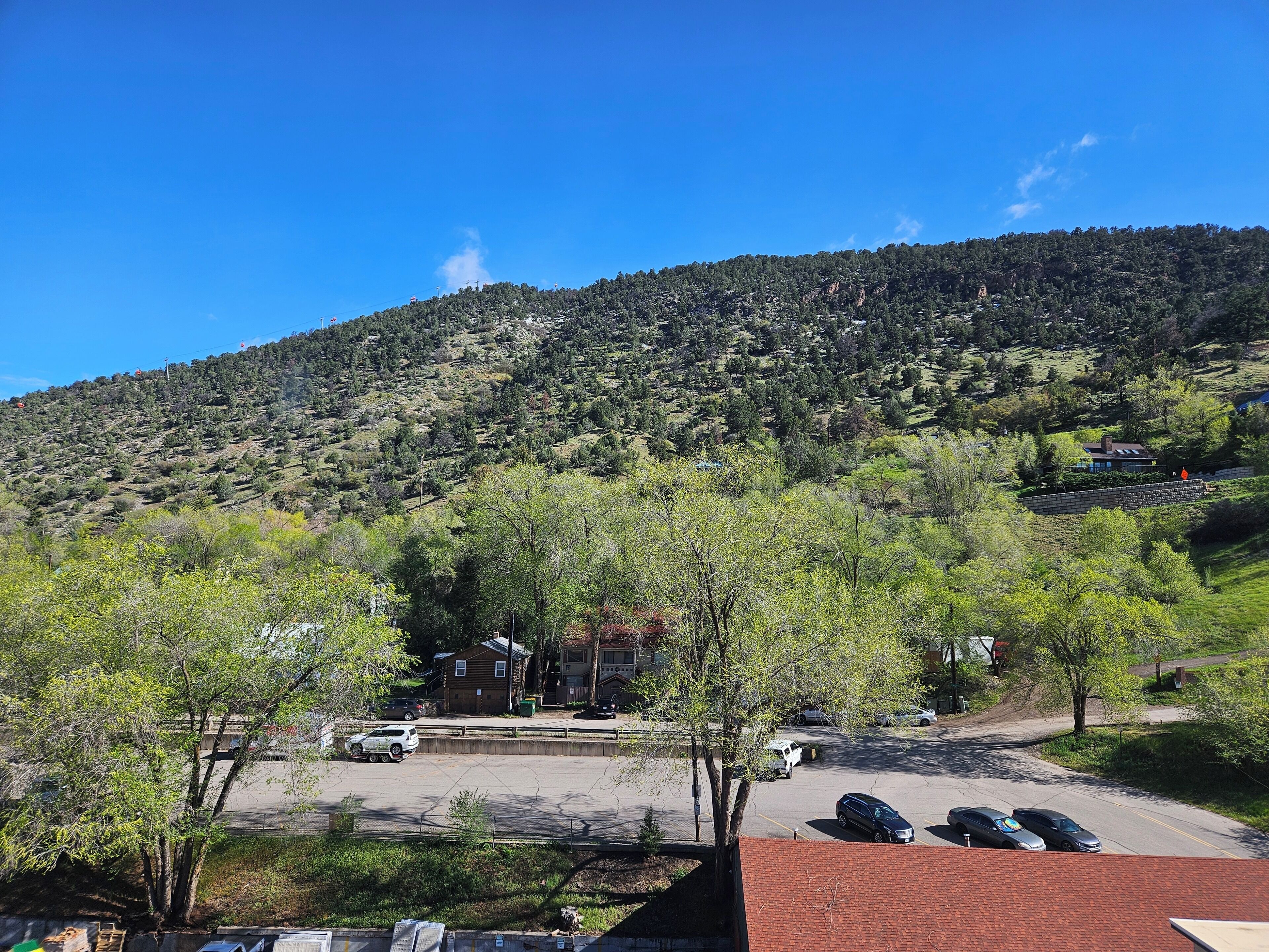 A parking area with several cars, a building with a red roof, and a hill with trees in the background.