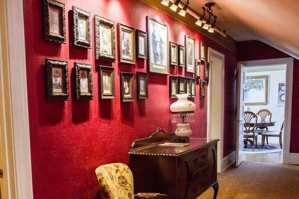 A hallway with a red wall adorned with framed pictures, a wooden chair, a desk with a lamp, and a door leading to another room.