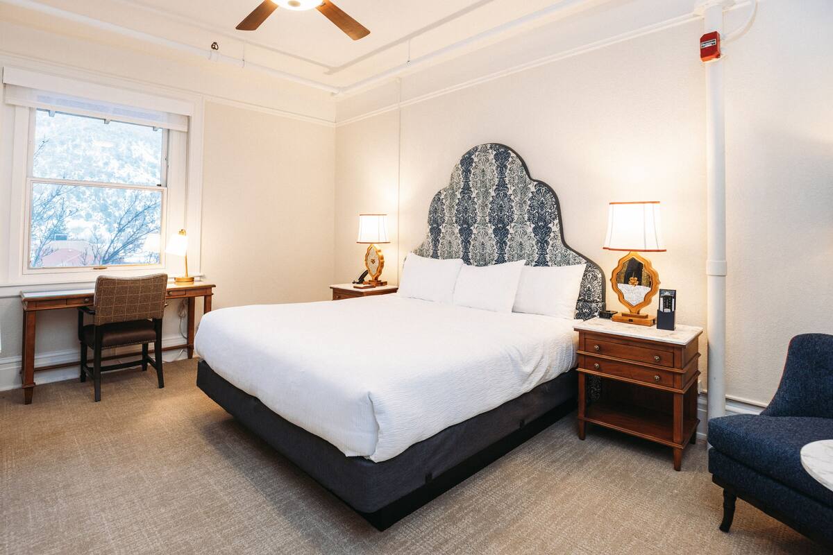 A hotel room with a large bed, two bedside tables with lamps, a desk, a chair, and a view of trees through the window.