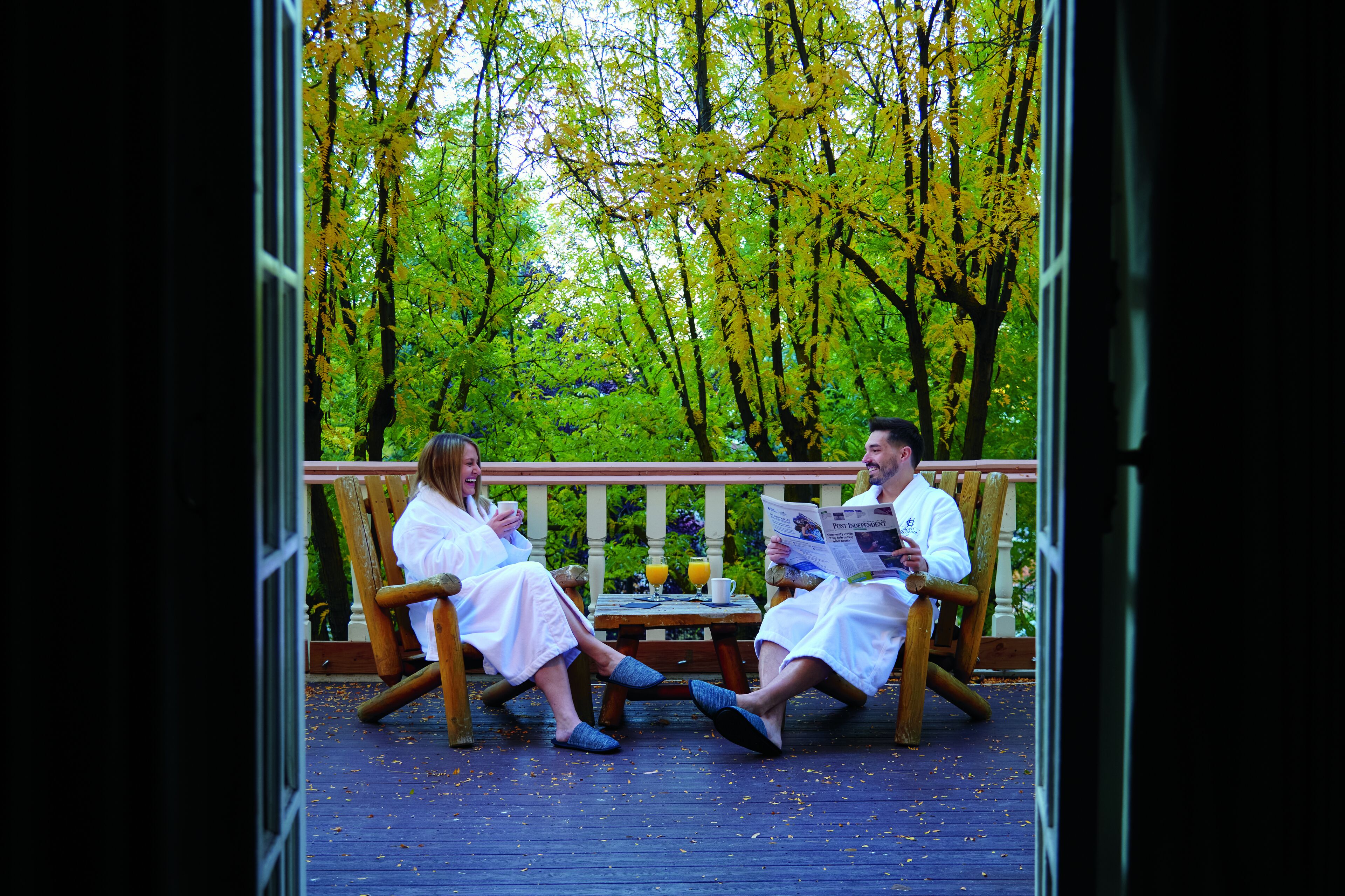 Two people sitting on wooden chairs on a wooden deck, reading a newspaper.