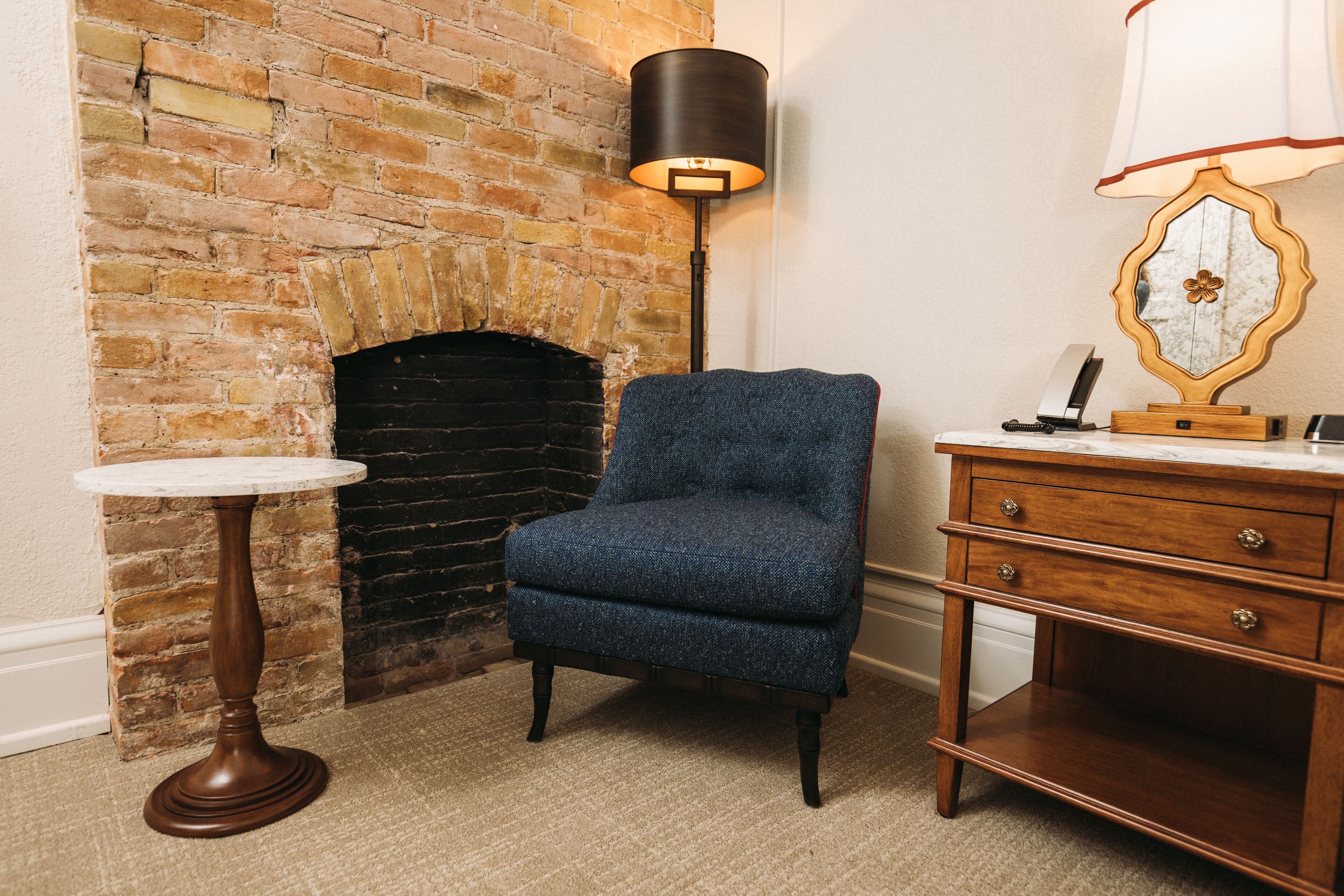 Una habitación con chimenea de ladrillo, un sillón azul, una mesa redonda de madera, una lámpara de pie y un aparador de madera con cajones.