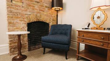 Una habitación con chimenea de ladrillo, un sillón azul, una mesa redonda de madera, una lámpara de pie y un aparador de madera con cajones.