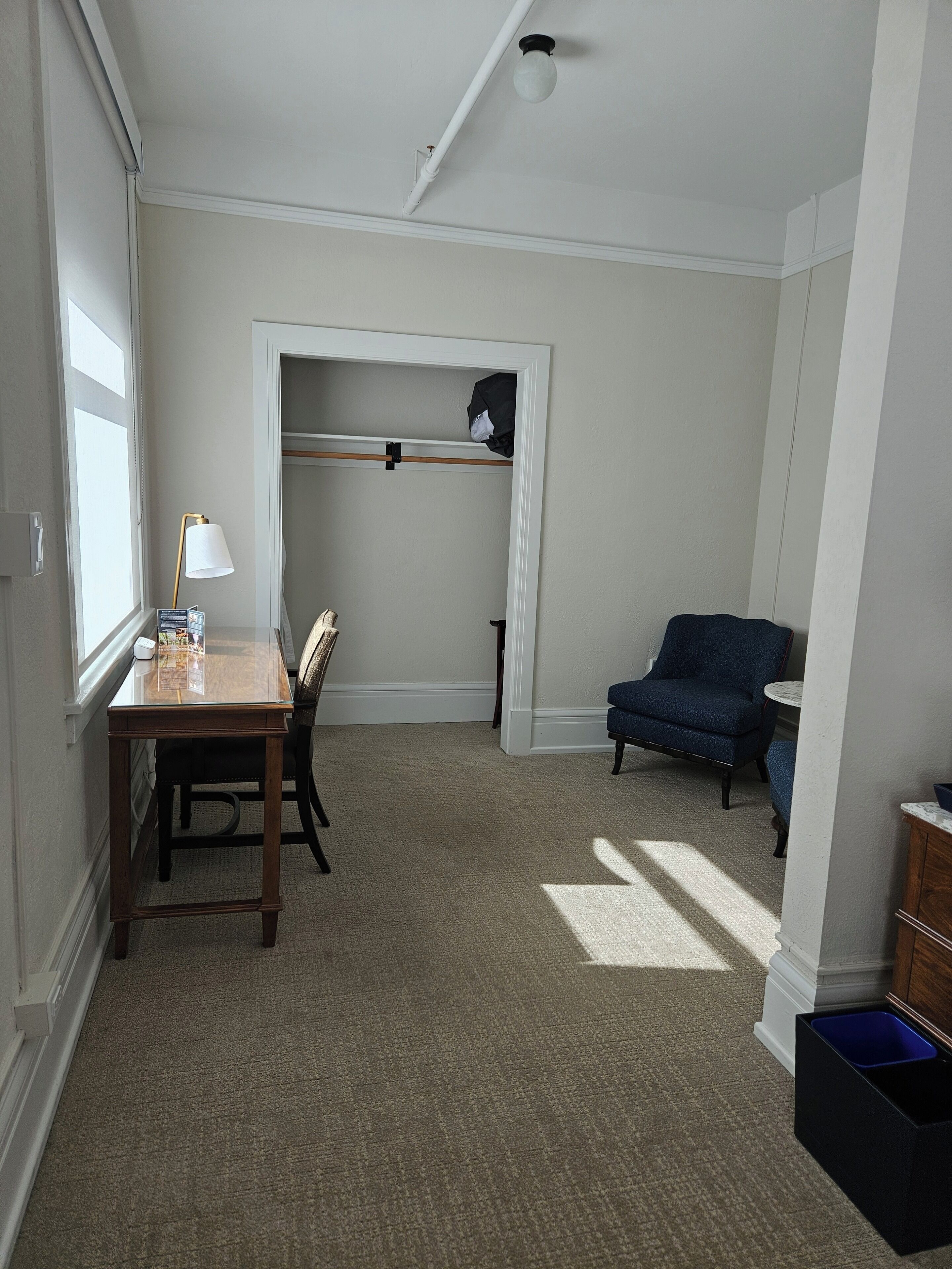 A room with a wooden desk, a chair, a blue armchair, a small table, and a closet with a coat rack.