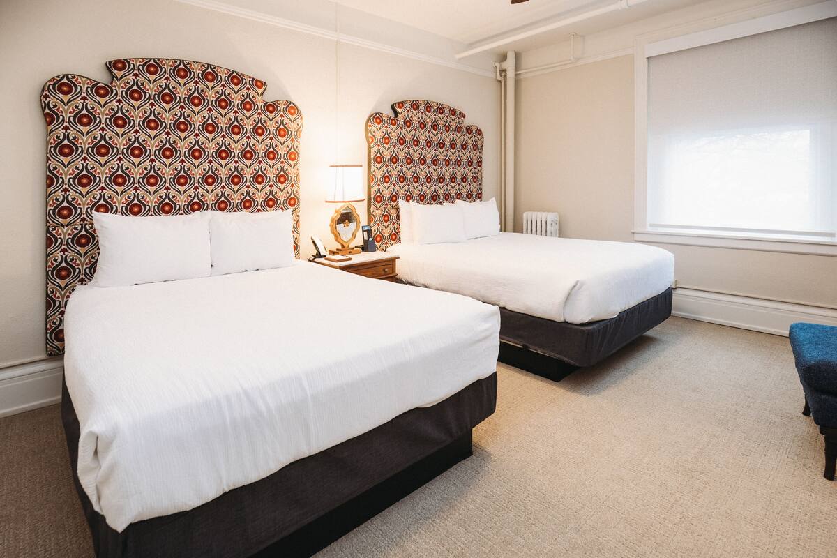 A hotel room with two beds, a nightstand with a lamp, a window with blinds, and a blue chair.