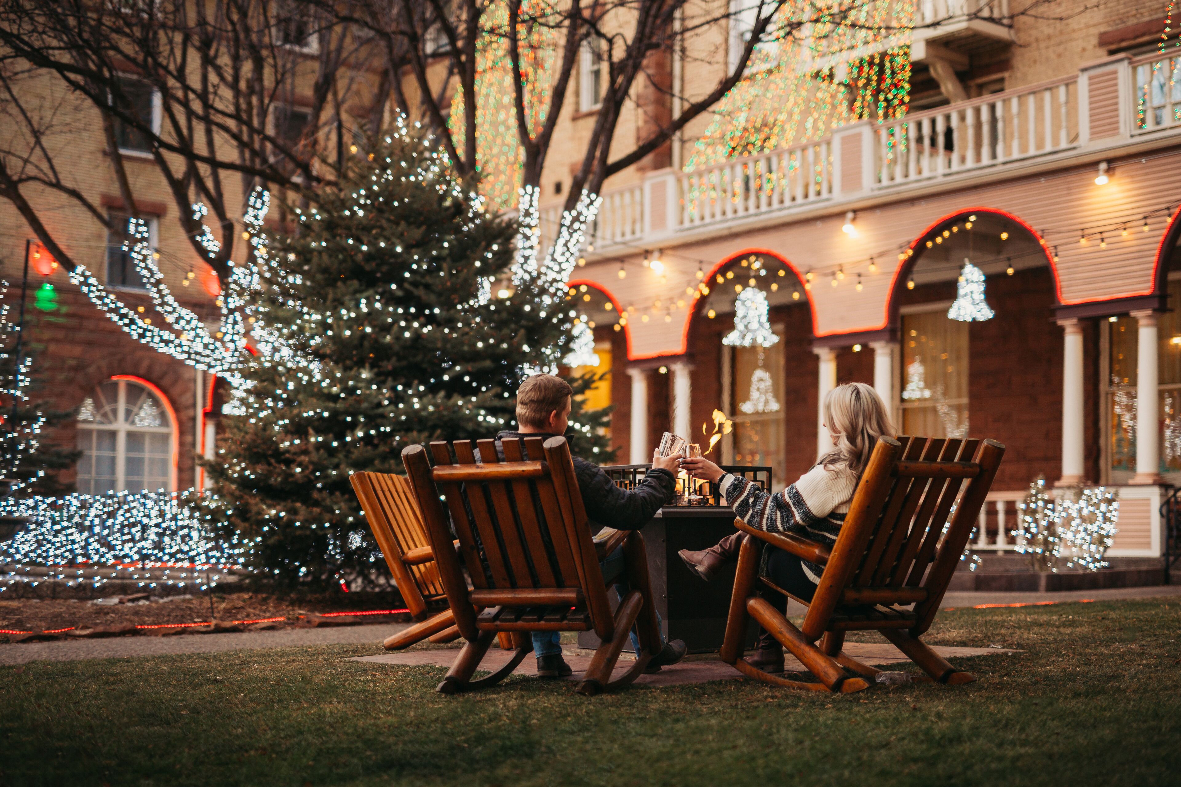 A cozy outdoor seating area with wooden chairs and a table, surrounded by a Christmas tree and festive lights.