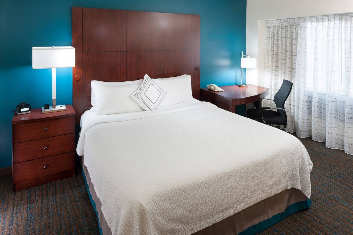 A hotel room with a large bed, a wooden headboard, a nightstand with a lamp, a desk with a chair, and a window with curtains.