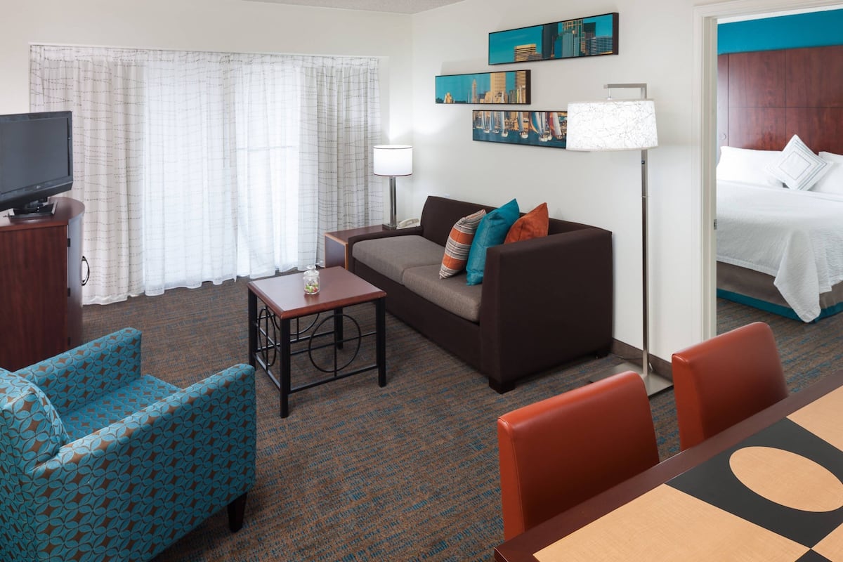 A hotel room with a sofa, armchair, TV, lamp, paintings, and a bed.