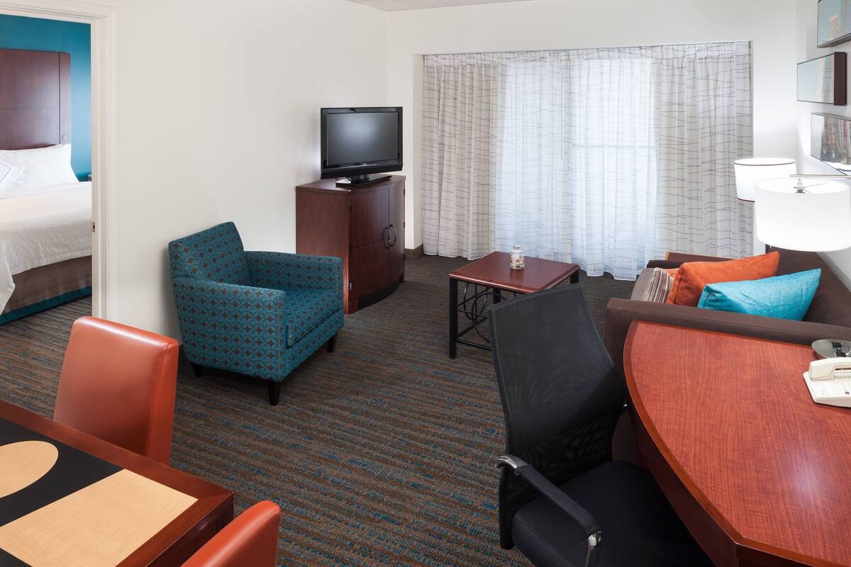 A hotel room with a bed, a TV on a wooden cabinet, a blue patterned armchair, a brown sofa with orange and blue cushions, a small table with a lamp, and a window with curtains.