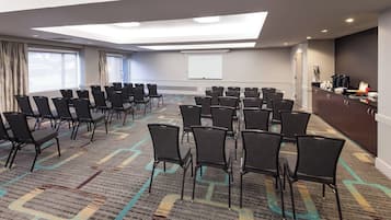Un salón de conferencias con filas de sillas, una pantalla de proyector y una zona de kitchenette.
