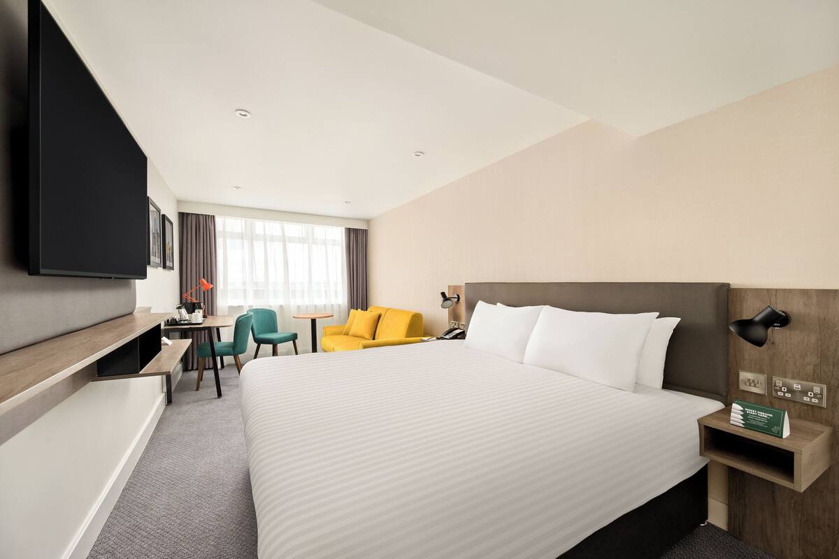 A hotel room with a large bed, a desk with a chair, a yellow armchair, and a TV mounted on the wall.
