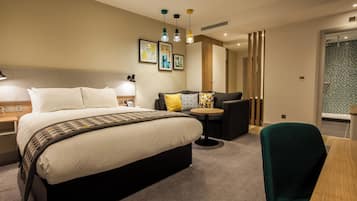 Une chambre d’hôtel moderne avec un grand lit, un canapé, une petite table et une chaise. On y trouve des lampes fixées au mur et une fenêtre avec vue.