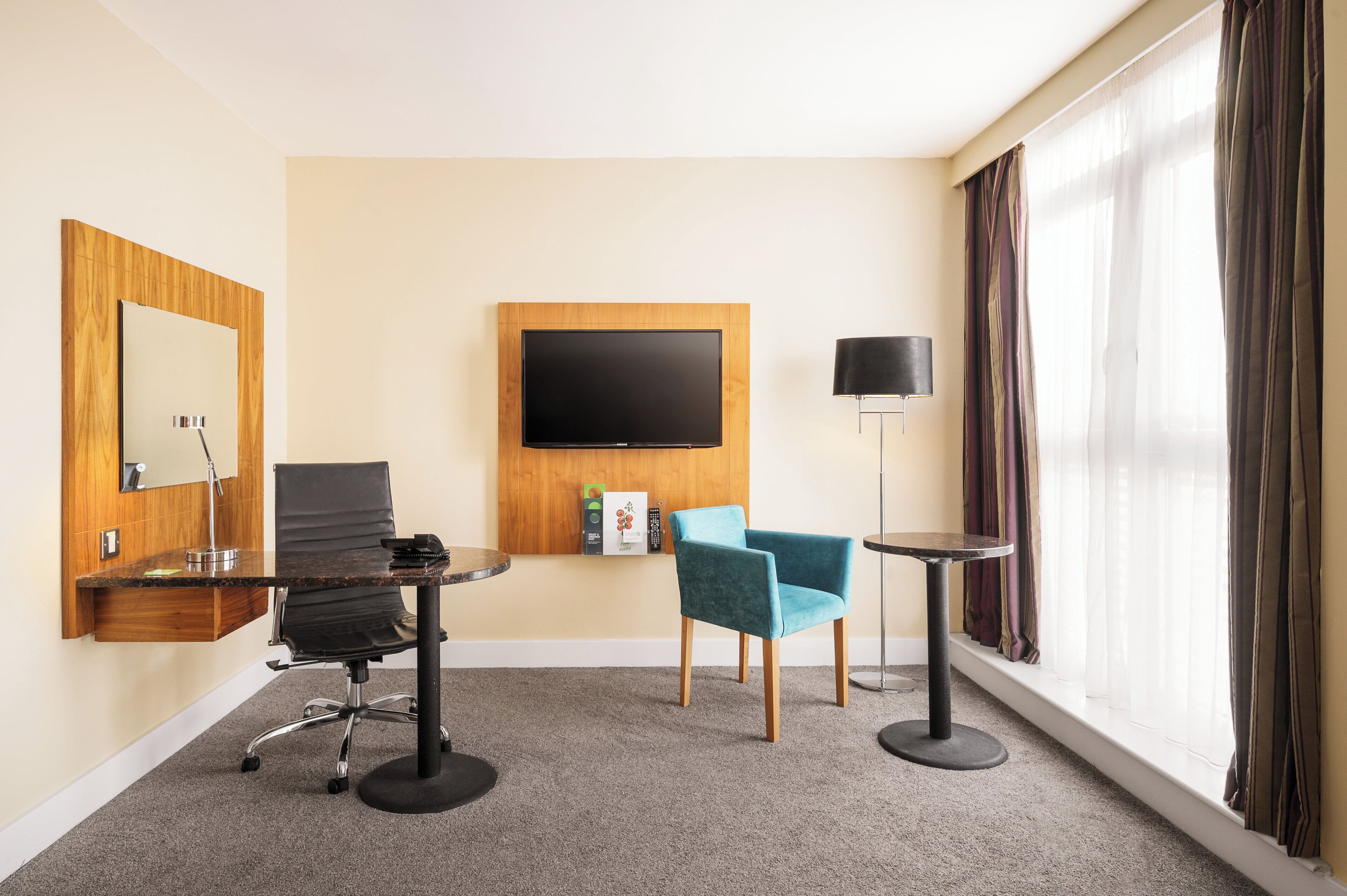 Ein Hotelzimmer mit einem an der Wand befestigten Flachbildfernseher, einem Schreibtisch mit Stuhl, einem kleinen runden Tisch mit Lampe und einem großen Fenster mit Vorhängen.