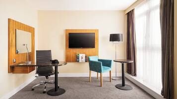 A hotel room with a flat-screen TV mounted on the wall, a desk with a chair, a small round table with a lamp, and a large window with curtains.