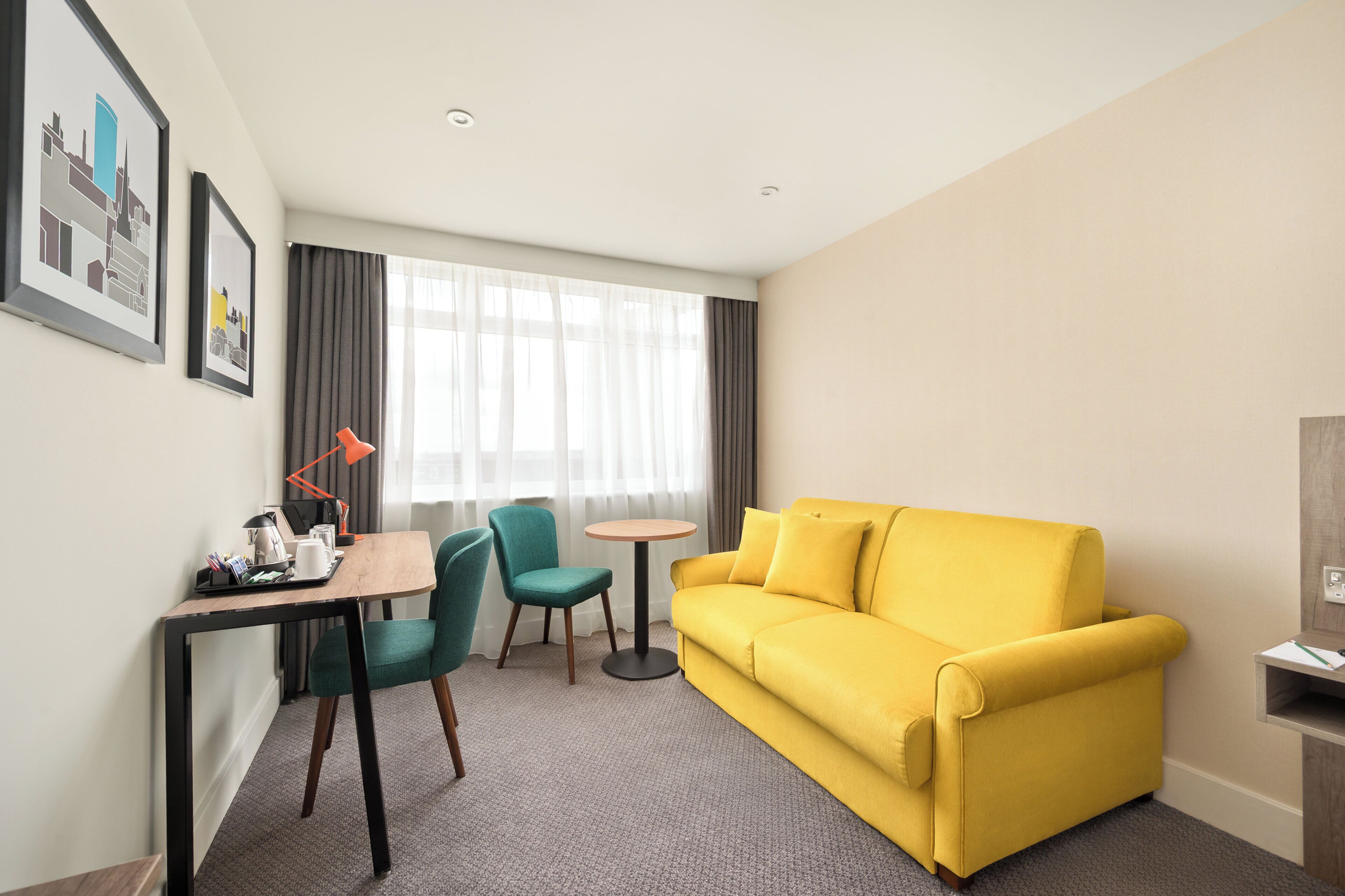 A hotel room with a yellow sofa, a desk with a lamp, two chairs, a small round table, and framed pictures on the wall.