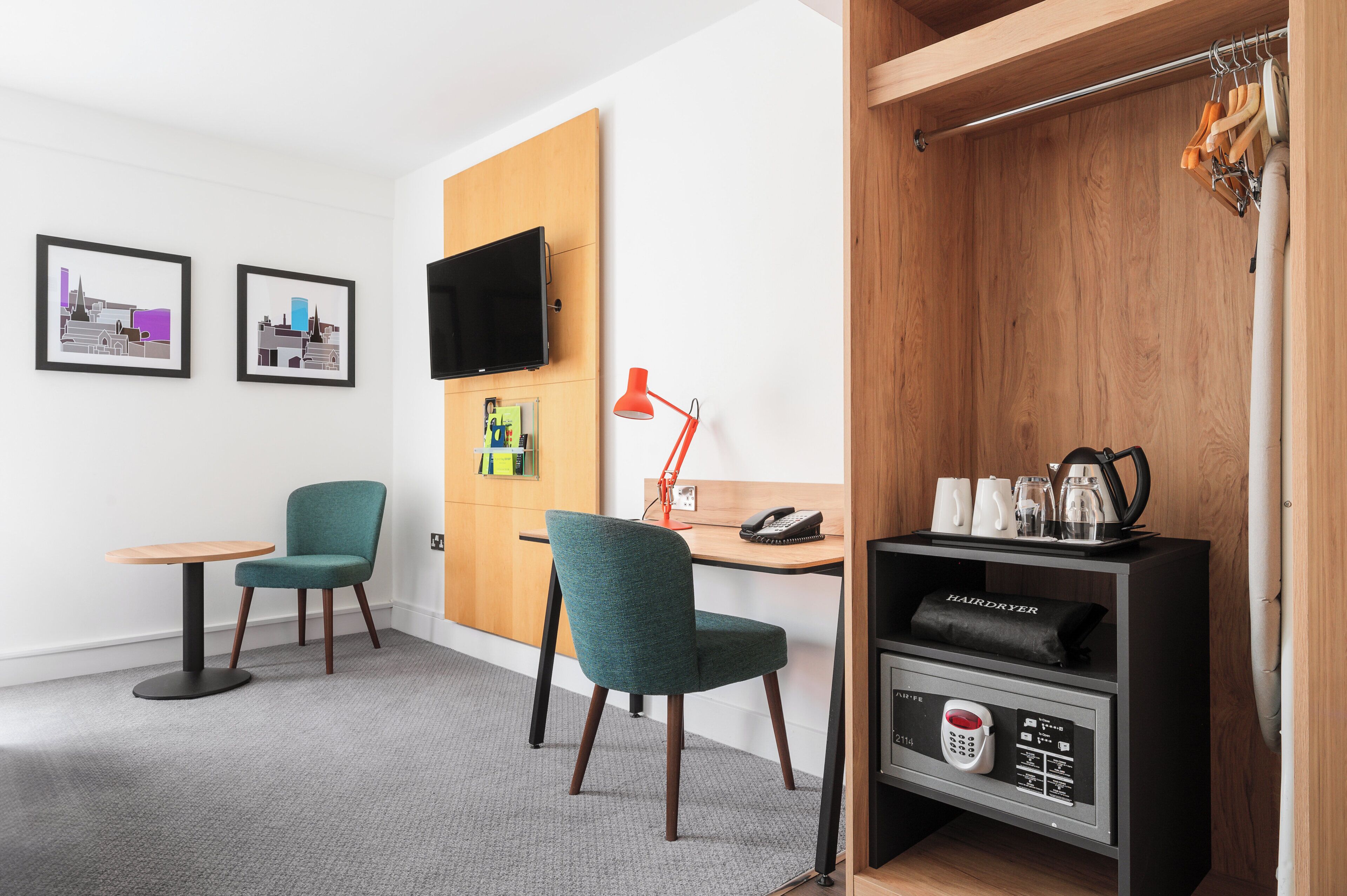 A hotel room with a wooden desk, two chairs, a TV mounted on the wall, a red desk lamp, and a hairdryer shelf.