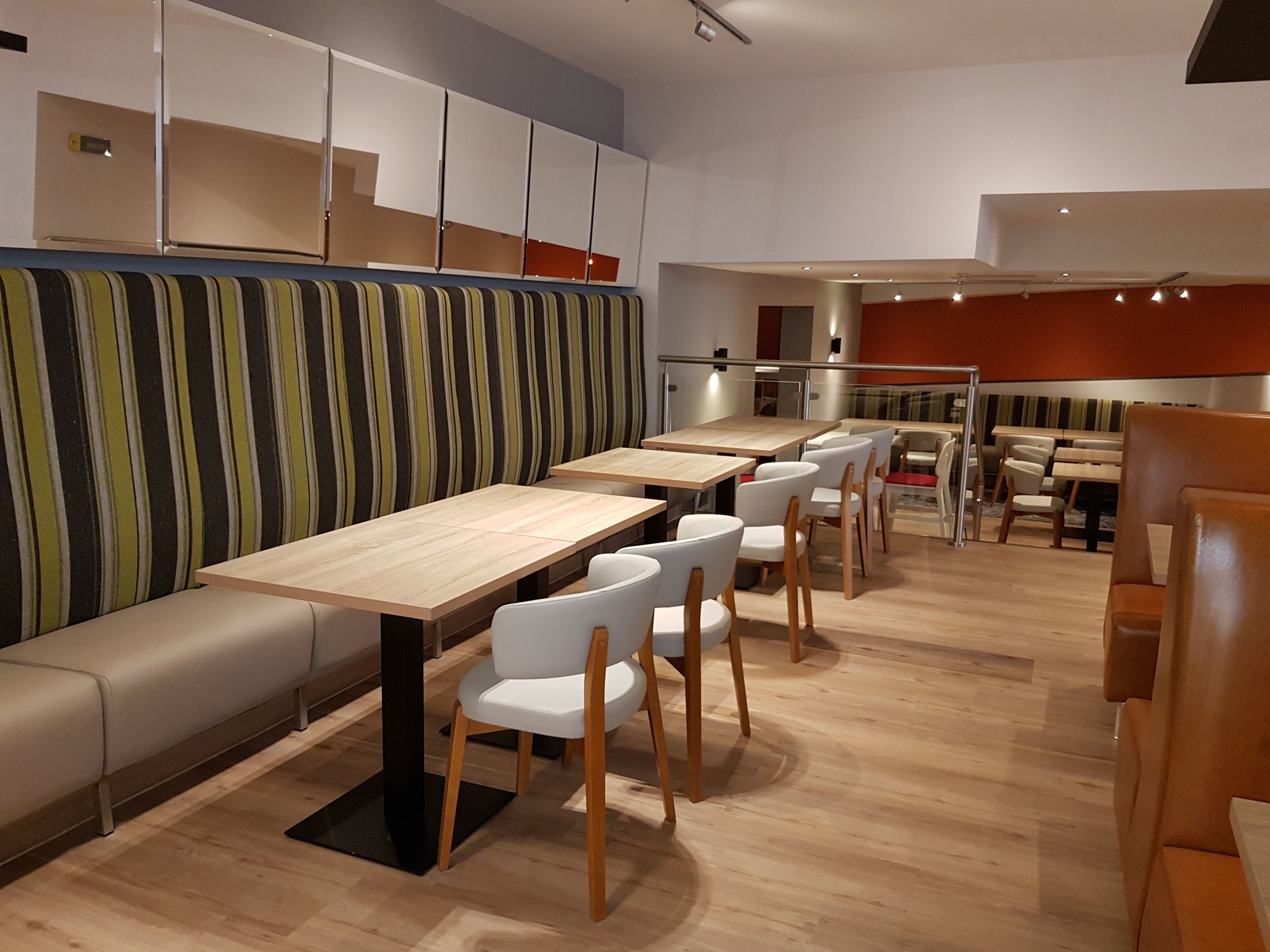 Un restaurante moderno con largas mesas de madera y sillas blancas, una pared a rayas y una zona de estar con bancos.