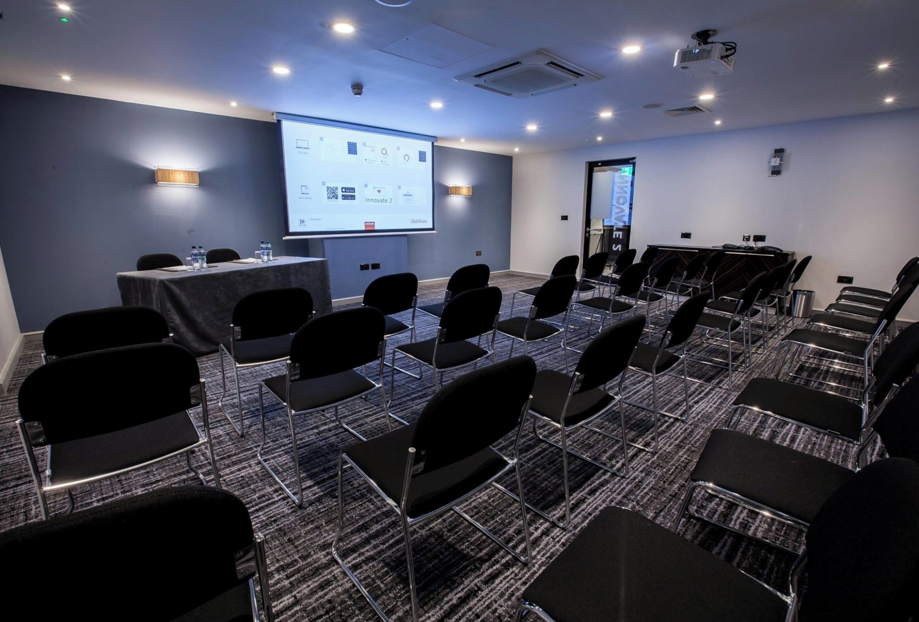 Un salón de conferencias con filas de sillas, una pantalla de proyección y una mesa con botellas de agua.