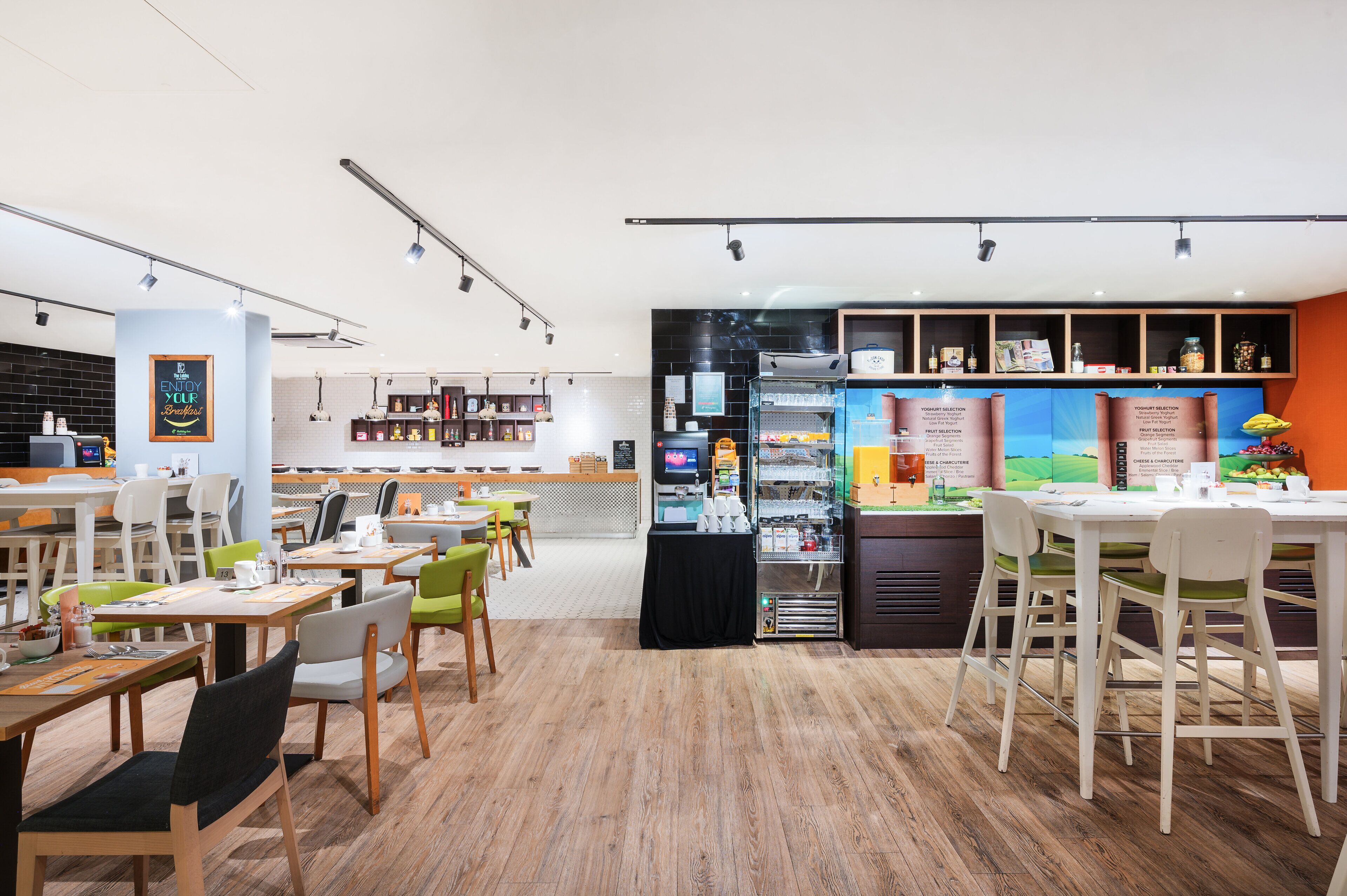 A modern café with wooden flooring, white and green chairs, and a counter displaying various food items.