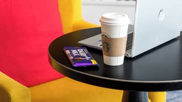 A yellow armchair with a red pillow, a black table with a laptop, a coffee cup, and a chocolate bar.
