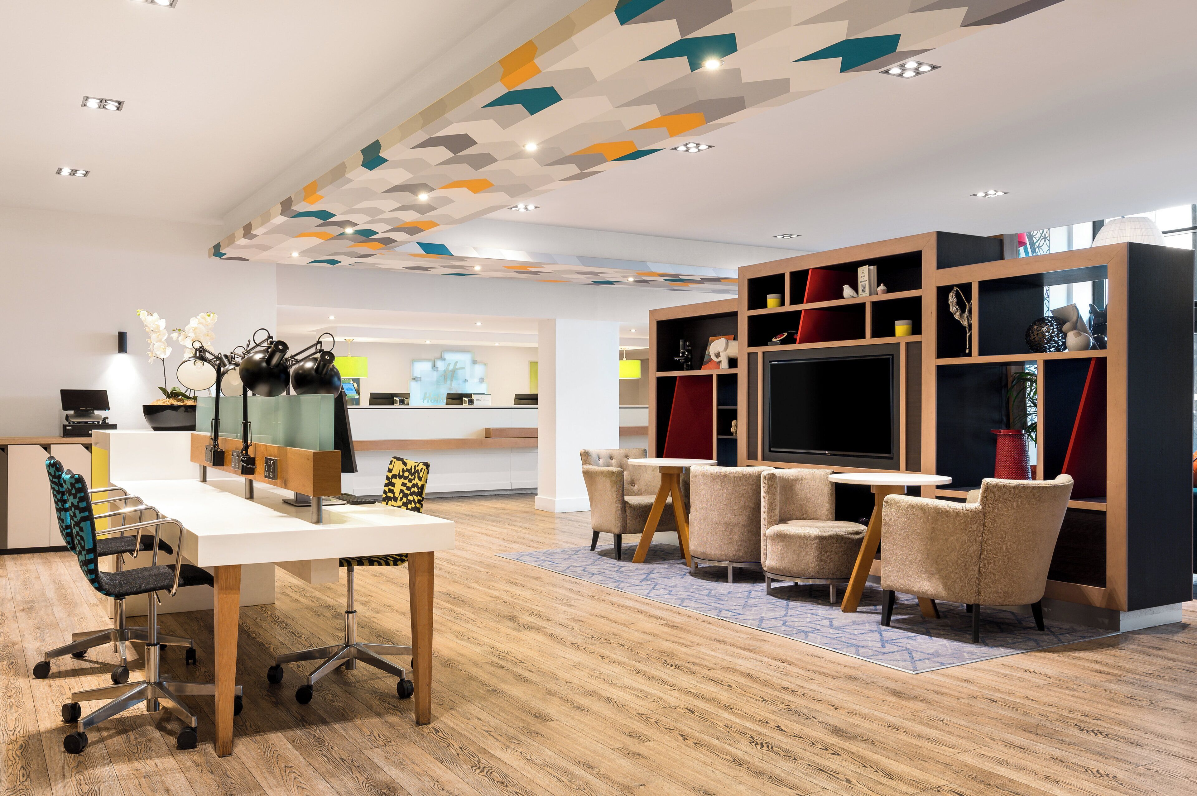 A modern lounge area with a white desk, ergonomic chairs, a large TV, and a bookshelf with various decorative items.