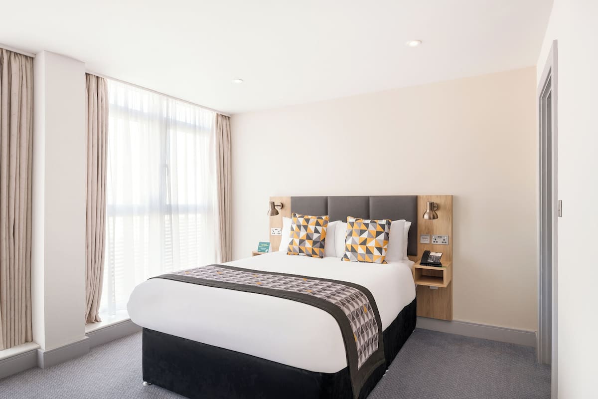 A hotel room with a large bed, a bedside table with a lamp, and a window with curtains.