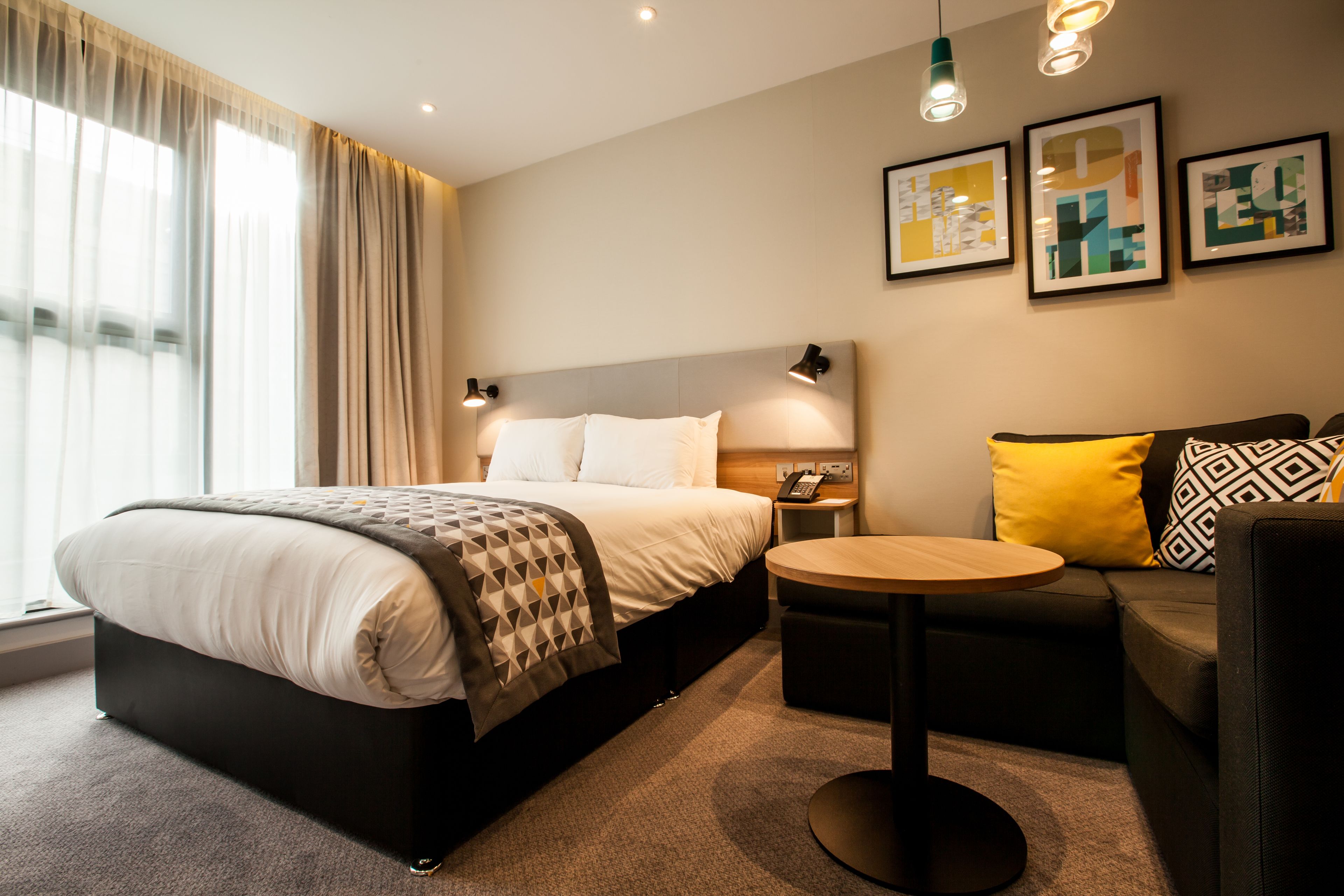 A modern hotel room with a large bed, a round table, a sofa, and artwork on the walls.