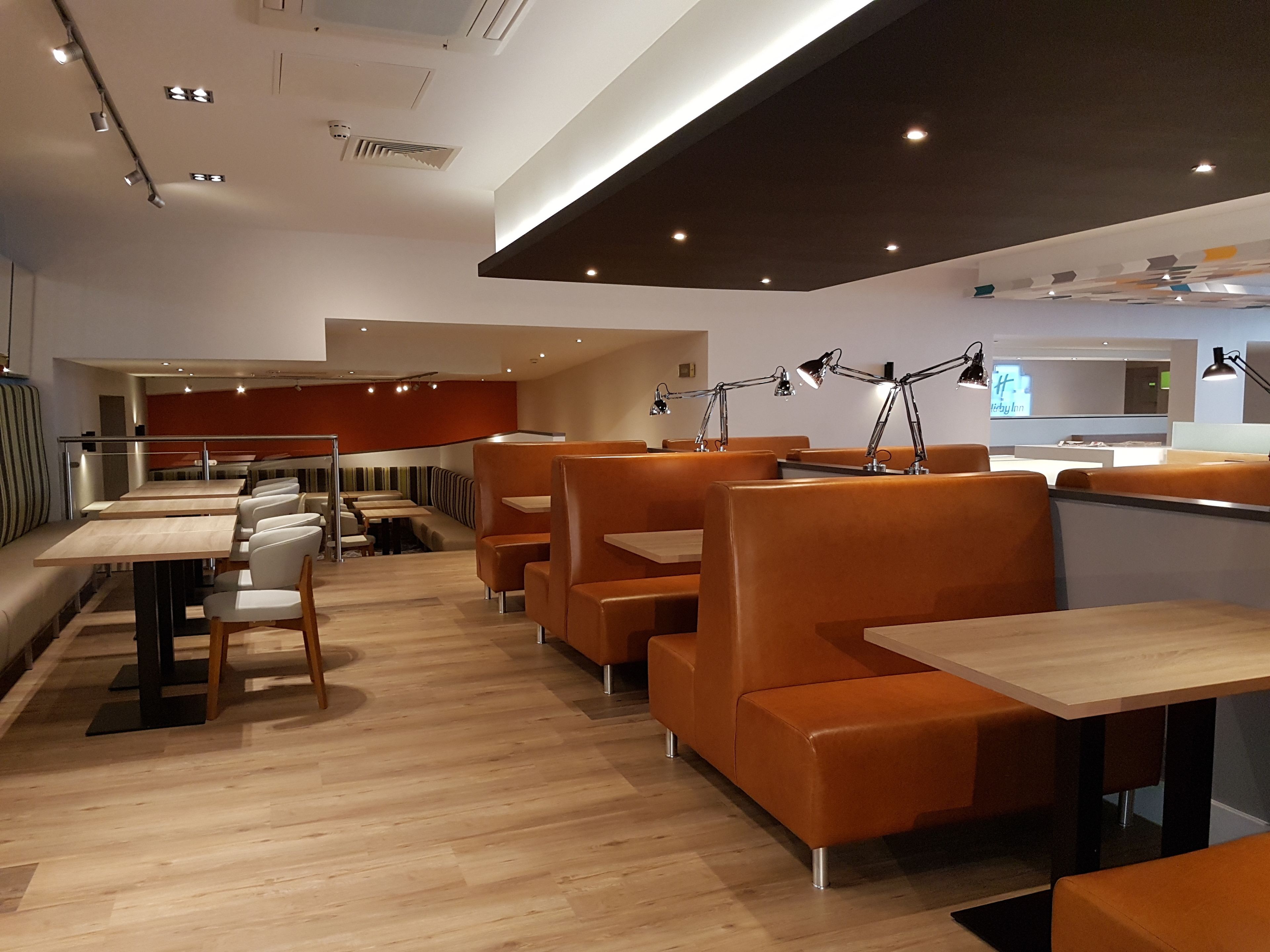 Ein modernes Restaurant mit Holztischen und orangefarbenen Sitzbänken, deckenintegraler Beleuchtung und einem sichtbaren Küchenbereich im Hintergrund.