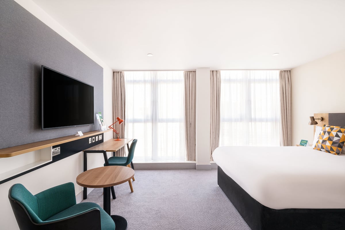 A hotel room with a bed, a desk with a chair, a TV, and large windows with curtains.