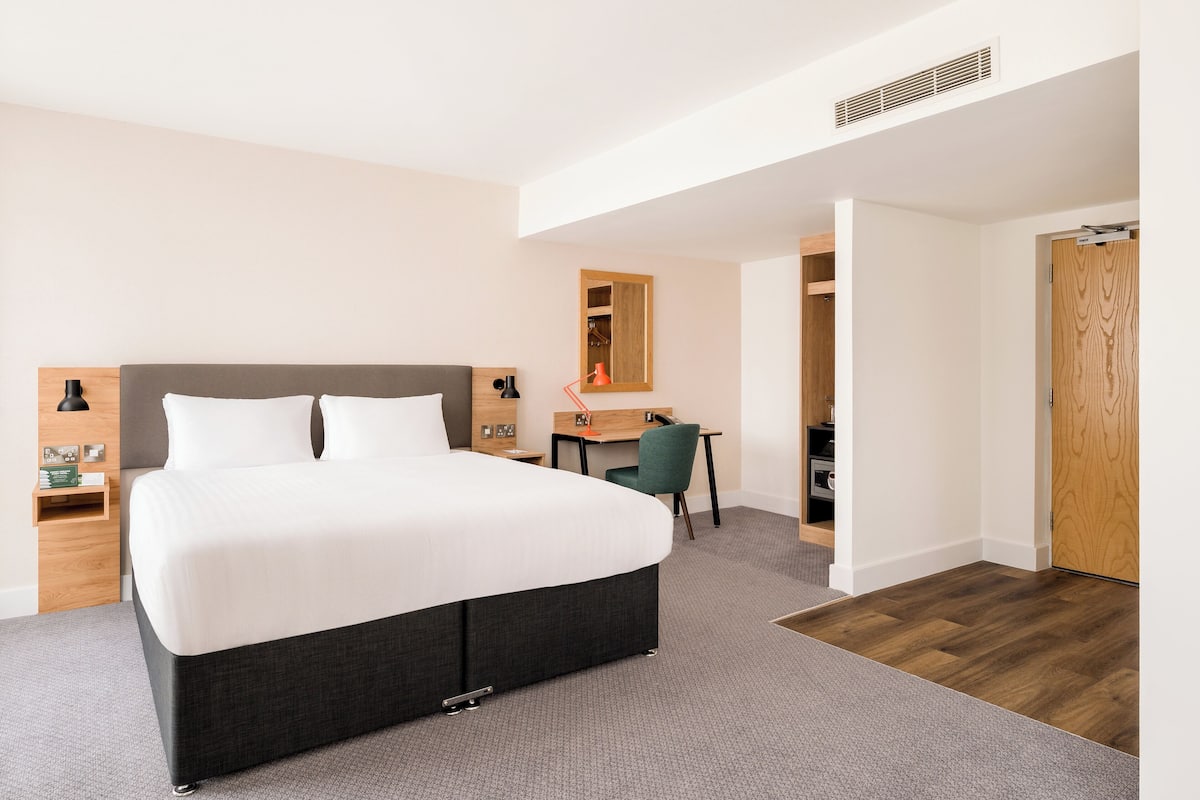 A modern hotel room with a large bed, a desk, a chair, and a wooden door.