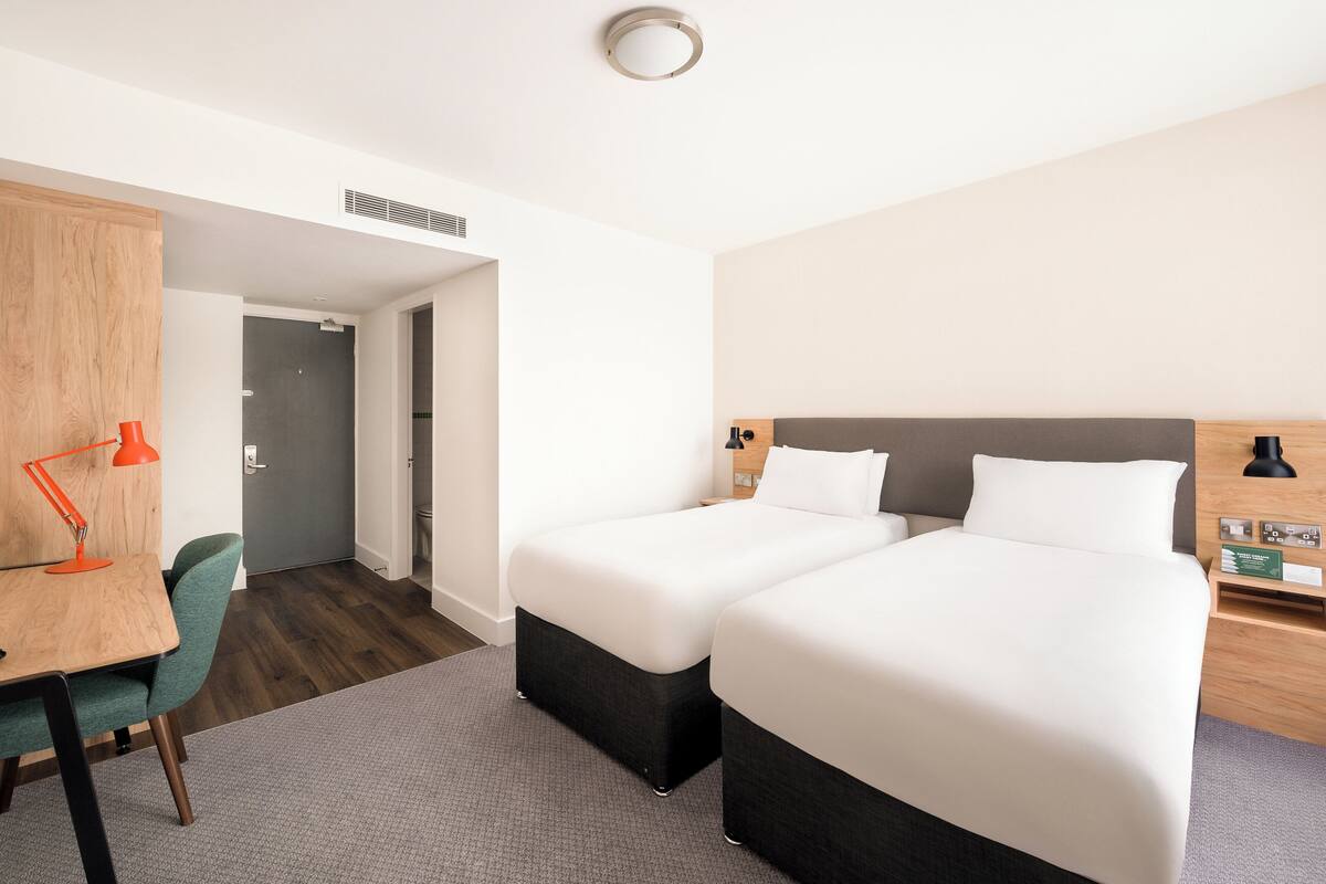 A hotel room with two beds, a desk with a chair, a lamp, and a wardrobe.