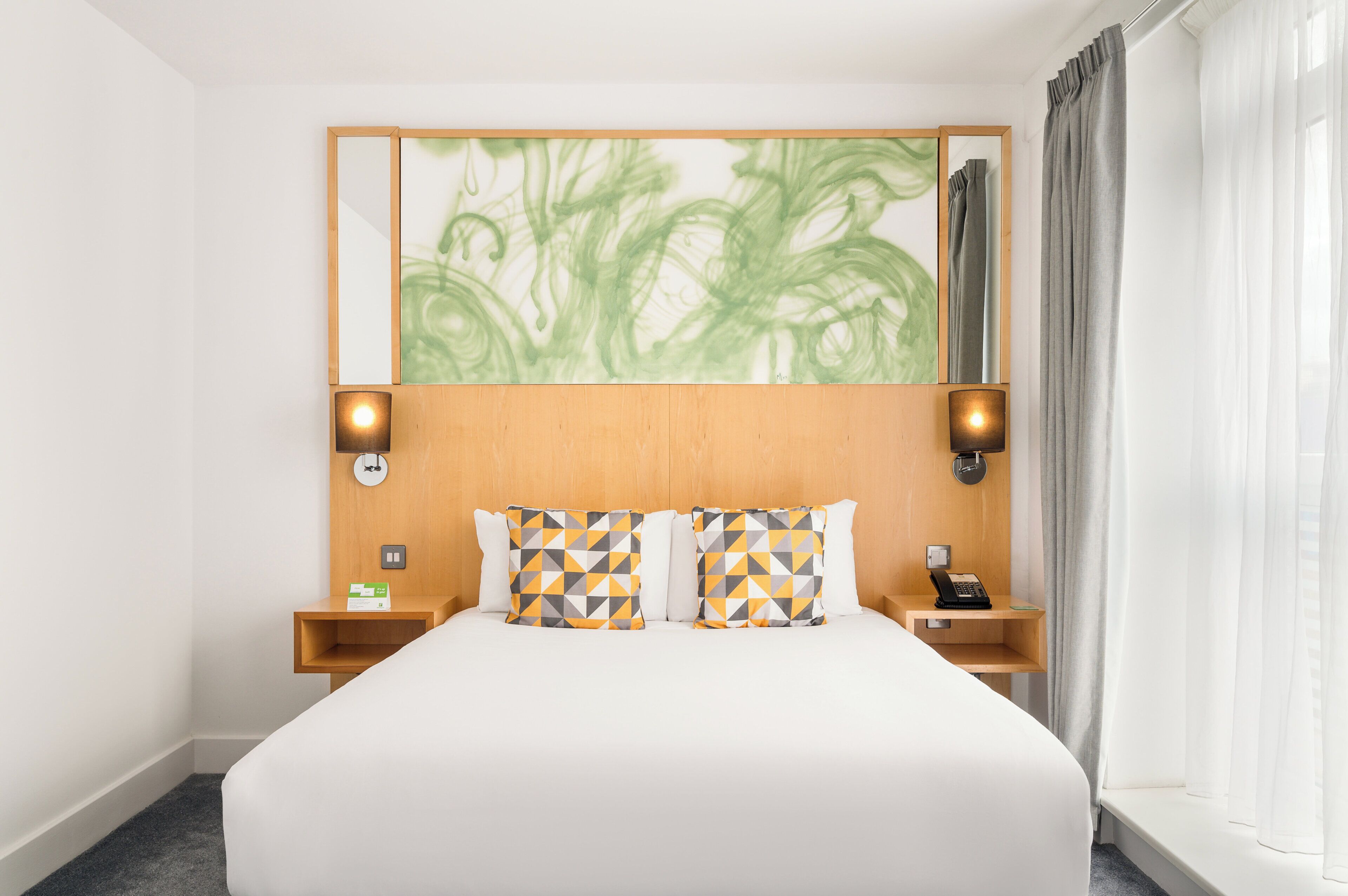 A hotel room with a large bed, two bedside tables, a telephone, and a mirror with a green abstract painting.