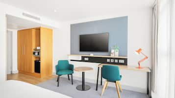 A modern hotel room with a wooden wardrobe, a flat-screen TV mounted on the wall, a desk with a computer and a lamp, and two teal chairs around a small round table.