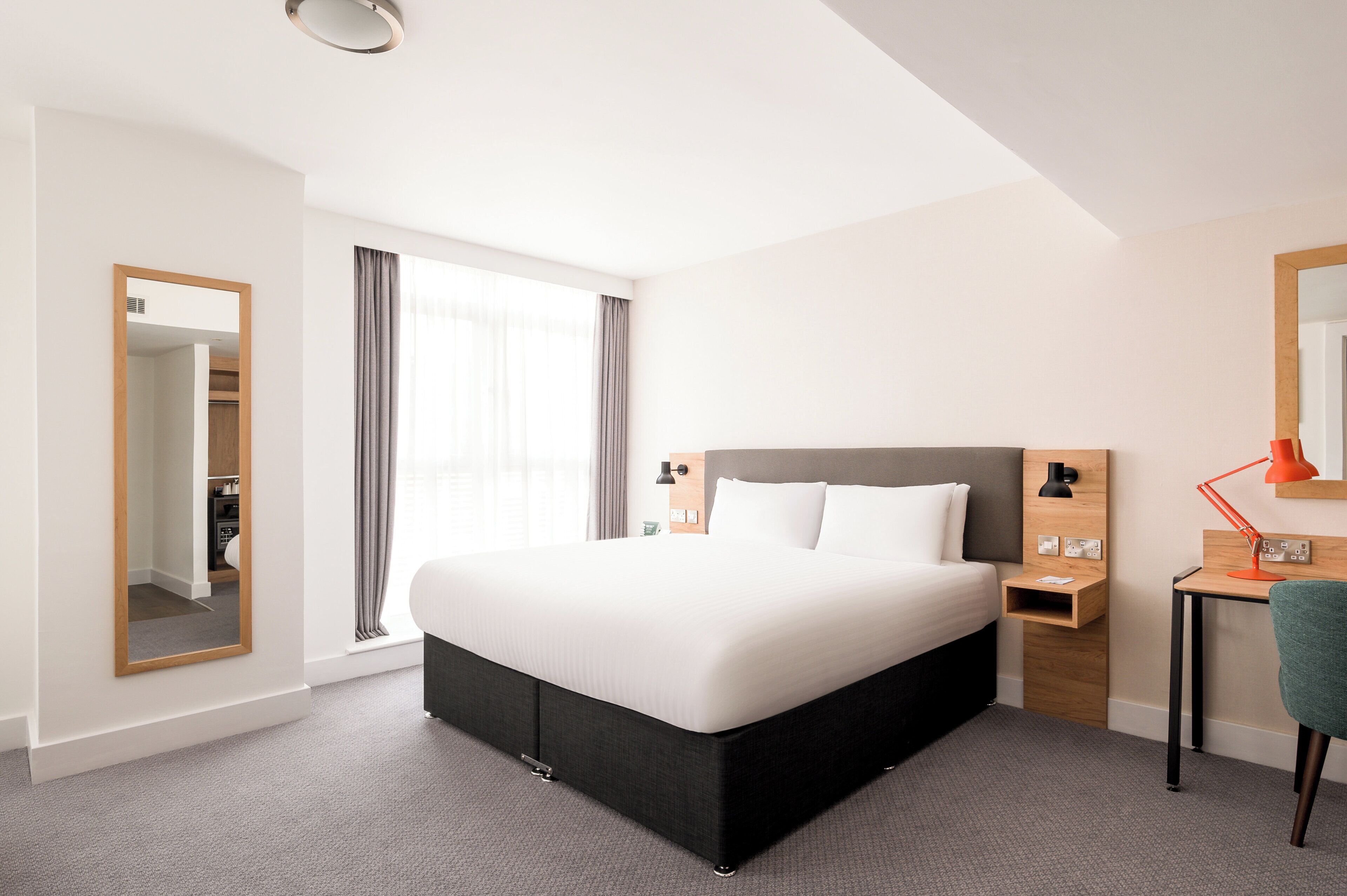 A modern hotel room with a large bed, a desk with a chair, a mirror, and a window with curtains.