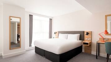 A modern hotel room with a large bed, a desk with a chair, a mirror, and a window with curtains.