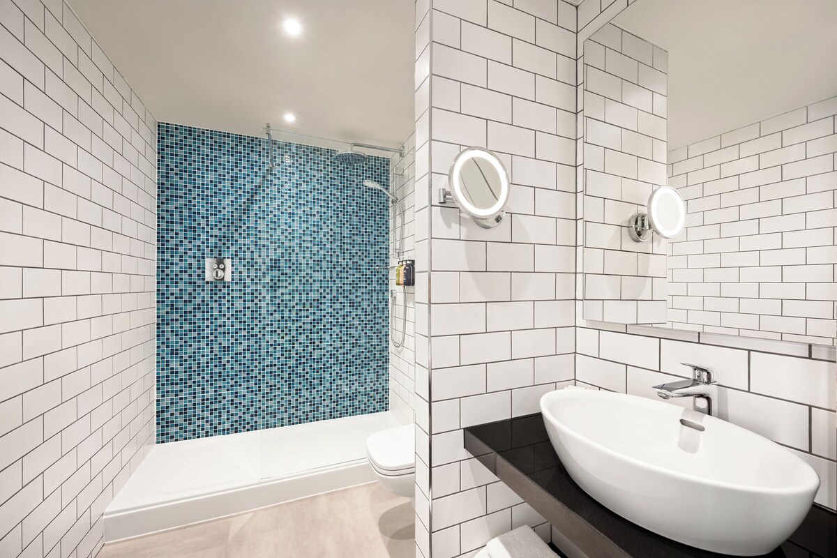 A modern bathroom with white tiled walls, a blue mosaic shower, a white toilet, and a black countertop with a white sink.